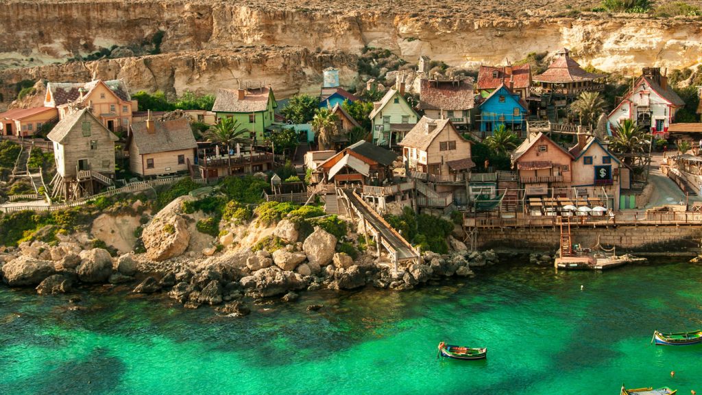 mellieha-malta Popeye Village em Mellieha, a melhor opção de cidade de praia onde ficar em Malta. As águas incrivelmente verdes são emolduradas pela silhueta dos barcos de pescadores, enquanto ao fundo, as rochas acompanham o curso do mar, destacando-se pelas pequenas e coloridas casas de madeira que pontuam a paisagem rochosa.