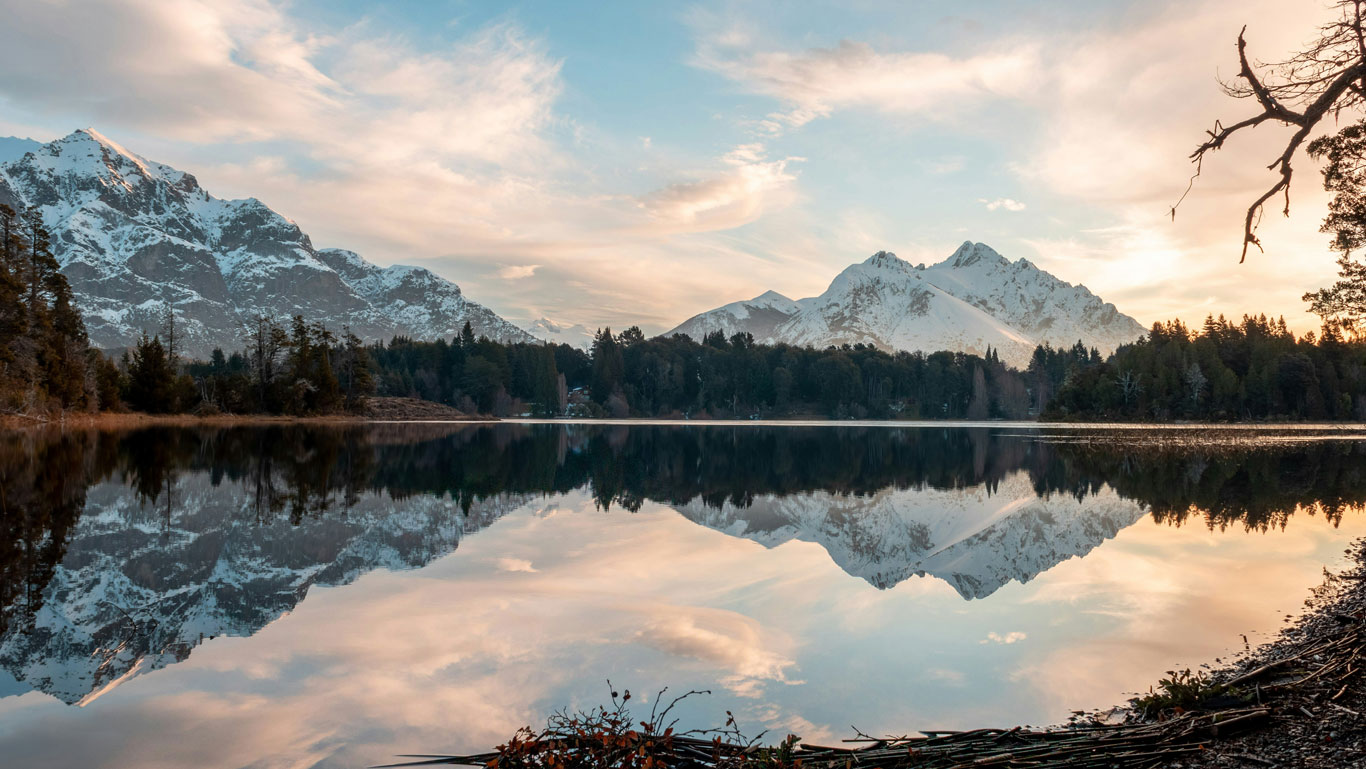 neve-em-bariloche Paisagem serena de um lago espelhado em Bariloche, Argentina, refletindo montanhas cobertas de neve em Bariloche ao entardecer. O céu apresenta tons suaves de azul e laranja, com nuvens leves, enquanto árvores e galhos secos emolduram a cena natural e tranquila.