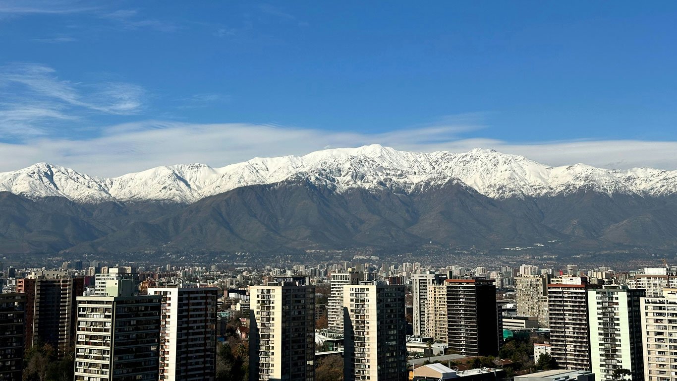 neve-em-santiago-cordilheira Skyline de Santiago com prédios altos em primeiro plano e a Cordilheira dos Andes coberta de neve ao fundo, sob um céu azul claro.