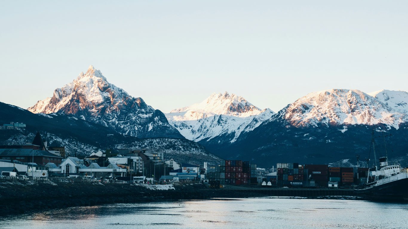neve-em-ushuaia Vista da cidade de Ushuaia com o porto e contêineres ao lado de um navio, tendo ao fundo montanhas com picos nevados iluminados pela luz do sol.