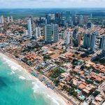 Vista aérea de Ponta Negra, o melhor bairro onde ficar em Natal. Com prédios modernos contrastando com as casas de praia, praias movimentadas e guarda-sóis coloridos alinhados na orla. O mar azul-turquesa contrasta com a urbanização intensa e o verde da vegetação ao fundo.