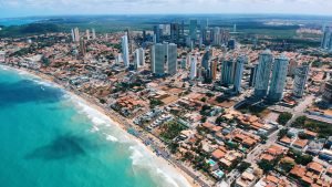 Vista aérea de Ponta Negra, o melhor bairro onde ficar em Natal. Com prédios modernos contrastando com as casas de praia, praias movimentadas e guarda-sóis coloridos alinhados na orla. O mar azul-turquesa contrasta com a urbanização intensa e o verde da vegetação ao fundo.