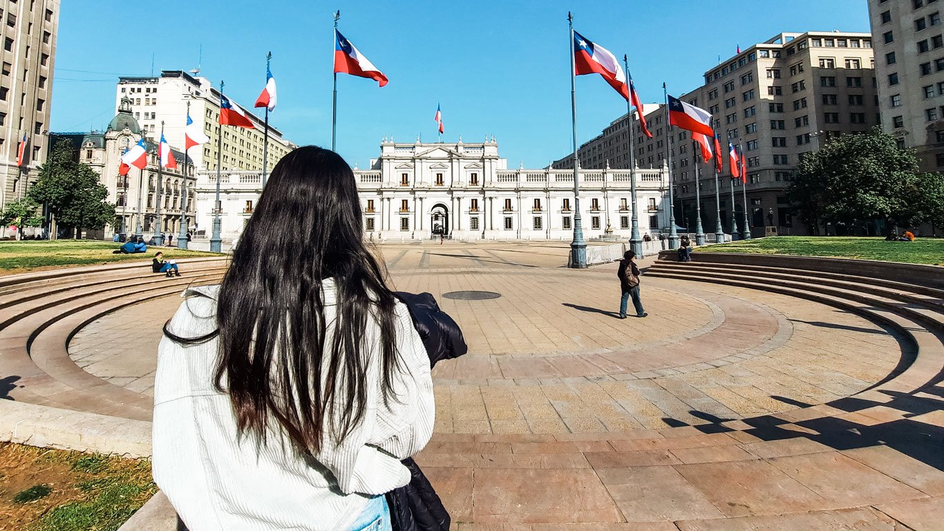 palacio-de-la-moneda-santiago Mulher de cabelos longos observa o Palácio de La Moneda, com bandeiras do Chile ao redor e céu limpo, em um típico dia de inverno na cidade.