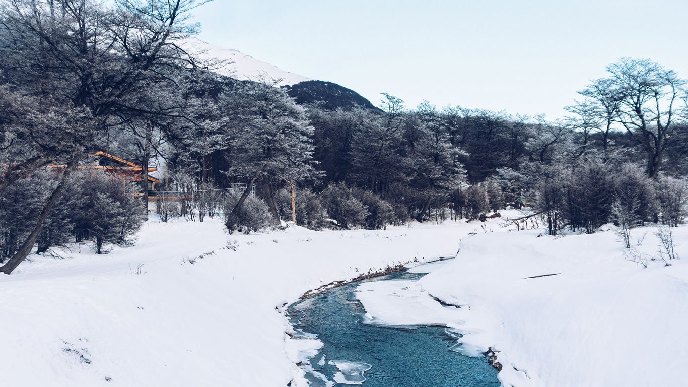 parque-nacional-tierra-del-fuego-ushuaia Riacho parcialmente congelado serpenteando por floresta coberta de neve no Parque Nacional Tierra del Fuego, com montanhas ao fundo.