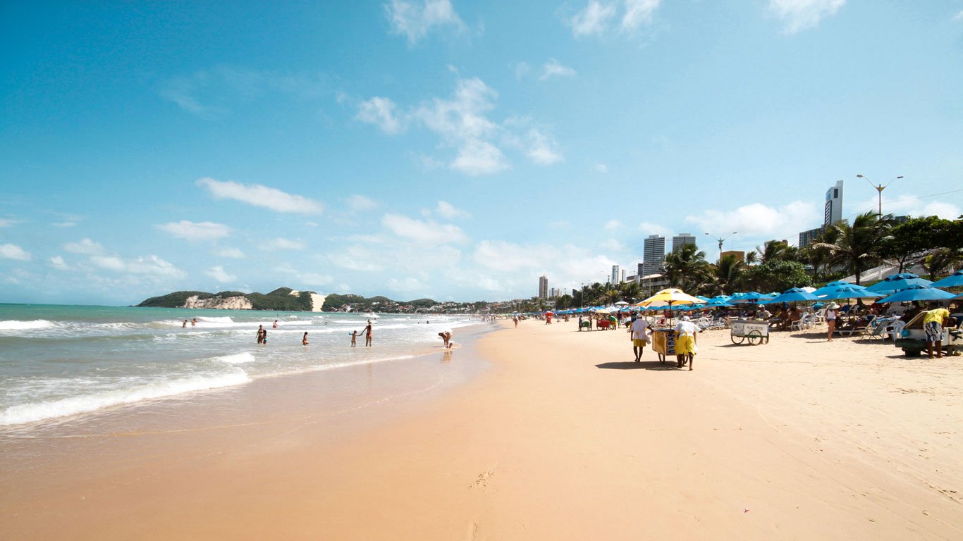 ponta-negra Praia de Ponta Negra com céu azul, pessoas se banhando no mar e vendedores ambulantes empurrando carrinhos sob guarda-sóis coloridos. Ao fundo, vê-se o Morro do Careca, famoso ponto turístico de Natal, RN.