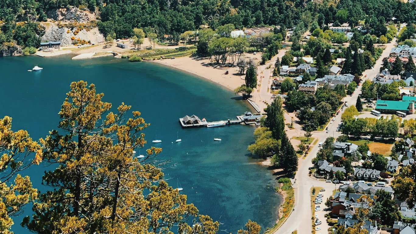 san-martin-de-los-andes Vista aérea de San Martín de los Andes, mostrando a orla do lago com um píer, pequenas embarcações ancoradas e uma praia de areia clara. A cidade aparece cercada por vegetação verde e montanhas, com ruas arborizadas e construções de estilo alpino.