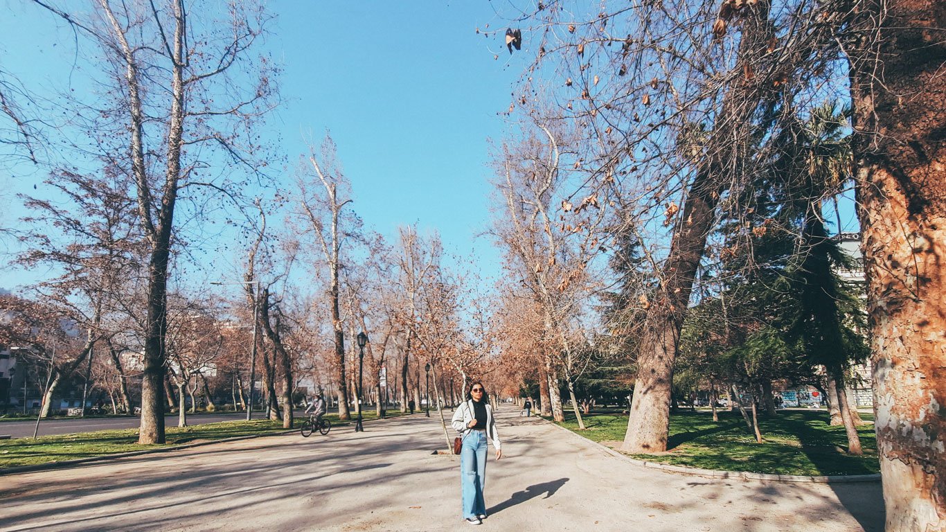 santiago-no-inverno-roupas Jovem caminha por uma alameda arborizada com folhas secas e árvores sem folhas no bairro Lastarria, cenário típico do inverno em Santiago.