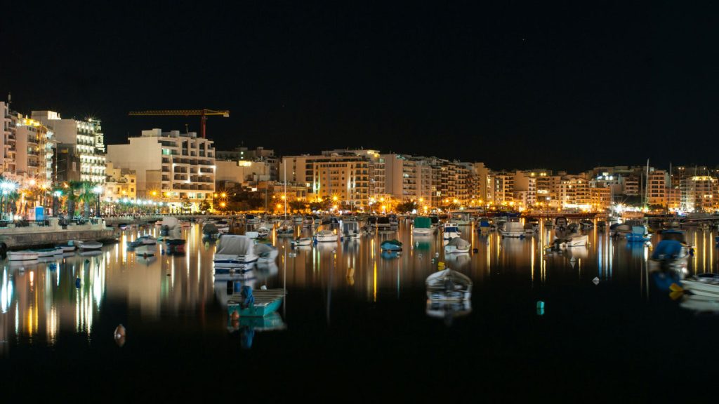 sliema-malta Baia de Sliema durante a noite, com as luzes da cidade refletindo no mar e iluminando os barcos ancorados, ao fundo os edifícios iluminados complementam a bela a paisagem.