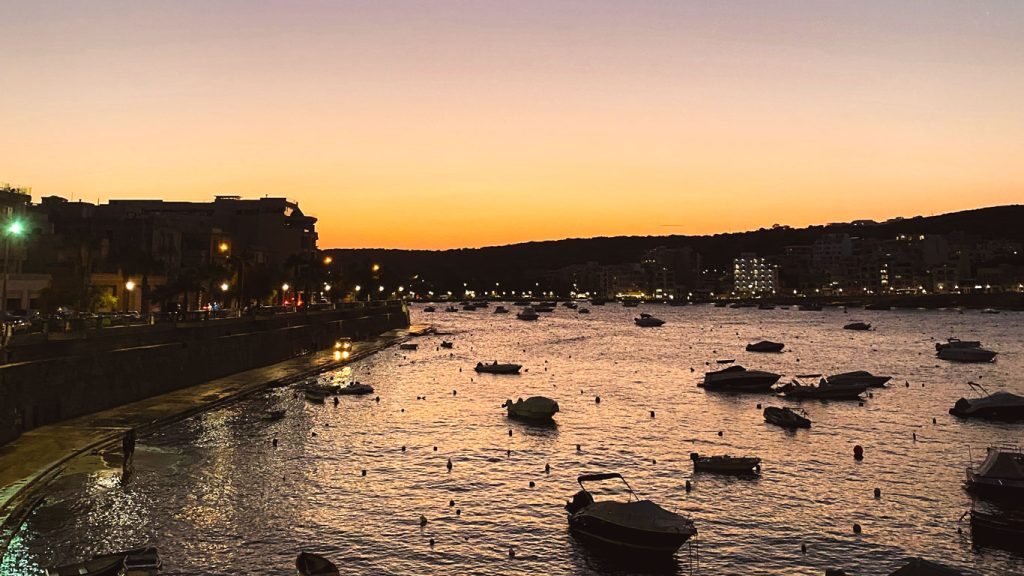 st-pauls-bay-malta St Paul’s Bay ao anoitecer, com os últimos raios do sol iluminando o mar e os pequenos barcos que estão ancorados. Ao lado esquerdo as luzes começam a se acender na orla que acompanha o mar.