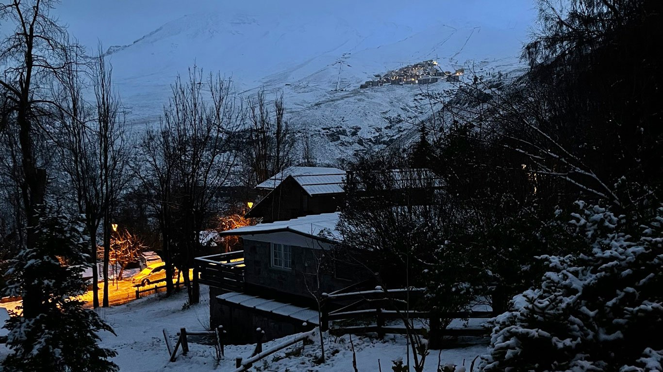 valle-nevado-chile Casas cobertas de neve ao entardecer no Valle Nevado em Santiago, com luzes amarelas iluminando a neve e vilarejo nas montanhas ao fundo parcialmente visível.