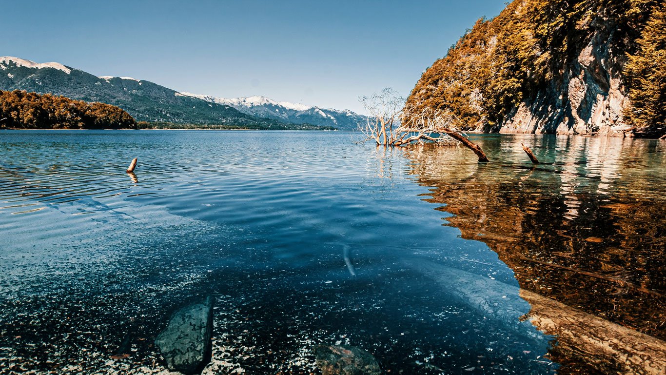 villa-la-angostura Imagem de um lago de águas cristalinas cercado por árvores e montanhas nevadas ao fundo, em Villa La Angostura. Galhos secos emergem da água e o reflexo da vegetação cria um efeito espelhado na superfície calma do lago.