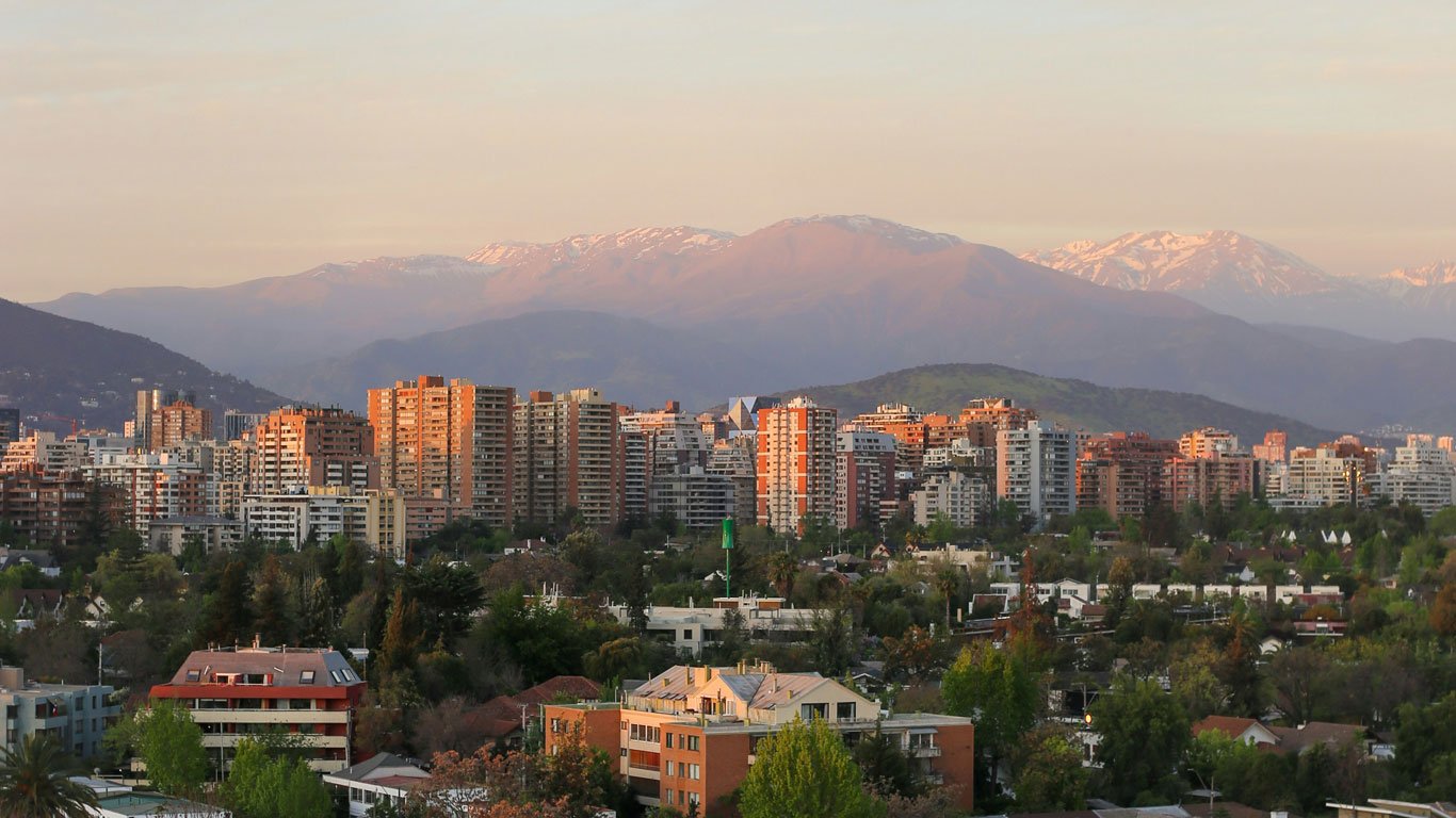 vista-para-a-cordilheira-dos-andes-em-santiago Vista panorâmica de Santiago ao entardecer com prédios altos e bairros arborizados em primeiro plano, tendo ao fundo a imponente Cordilheira dos Andes com picos nevados suavemente iluminados pela luz dourada do sol.