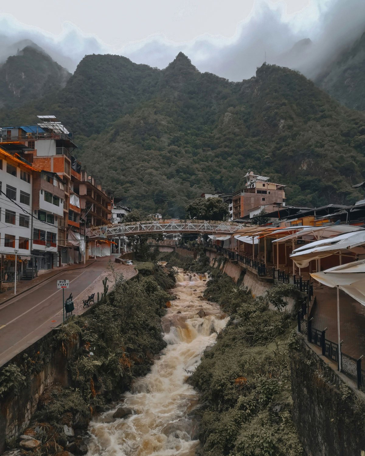 aguas-calientes-machu-picchu Vista do rio correndo entre construções de Águas Calientes, o melhor lugar onde ficar em Machu Picchu, cercado por montanhas cobertas de neblina.