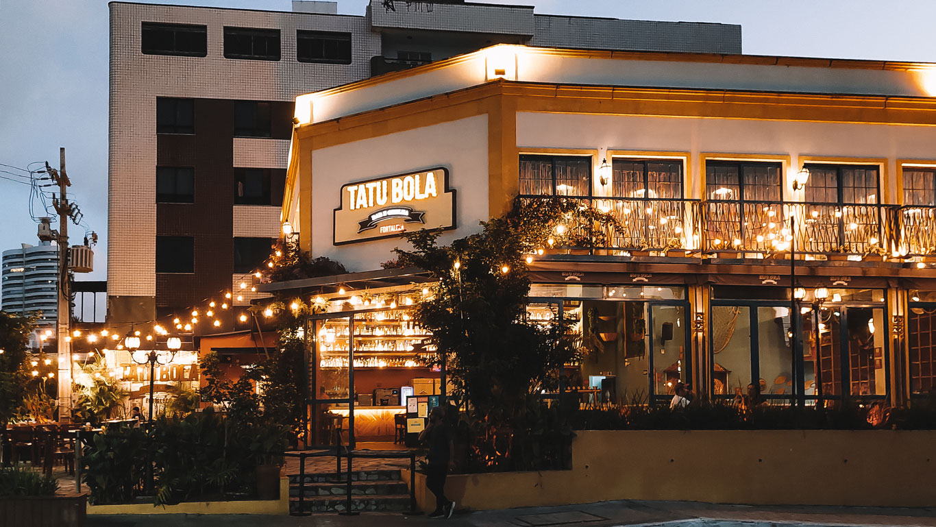 aldeota-fortaleza Fachada iluminada do bar Tatu Bola em Aldeota, Fortaleza, durante o anoitecer, com luzes decorativas criando um ambiente acolhedor e urbano, rodeado por prédios modernos.