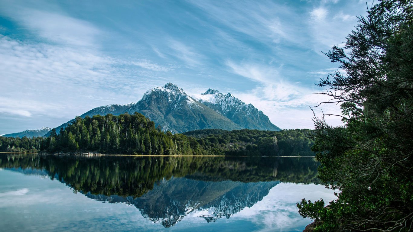 Montanhas nevadas em Bariloche, refletidas perfeitamente nas águas calmas de um lago, com céu azul e árvores densas na margem oposta.