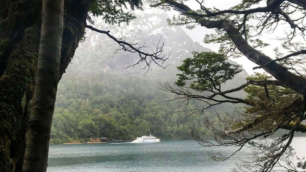 Vista de uma balsa branca ancorada no lago Nahuel Huapi em Bariloche em outubro. O lago está cercado por árvores verdes e montanhas cobertas de névoa ao fundo, com troncos e galhos emoldurando a cena.