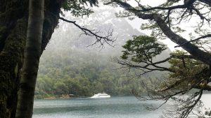 Vista de uma balsa branca ancorada no lago Nahuel Huapi em Bariloche em outubro. O lago está cercado por árvores verdes e montanhas cobertas de névoa ao fundo, com troncos e galhos emoldurando a cena.