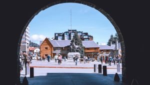 Praça central de Bariloche vista por um arco de pedra, com várias pessoas passeando com roupas de frio, casas de madeira e pedra ao fundo e uma escultura de cavalo no centro. O céu está limpo e azul, revelando um dia frio, porém ensolarado.