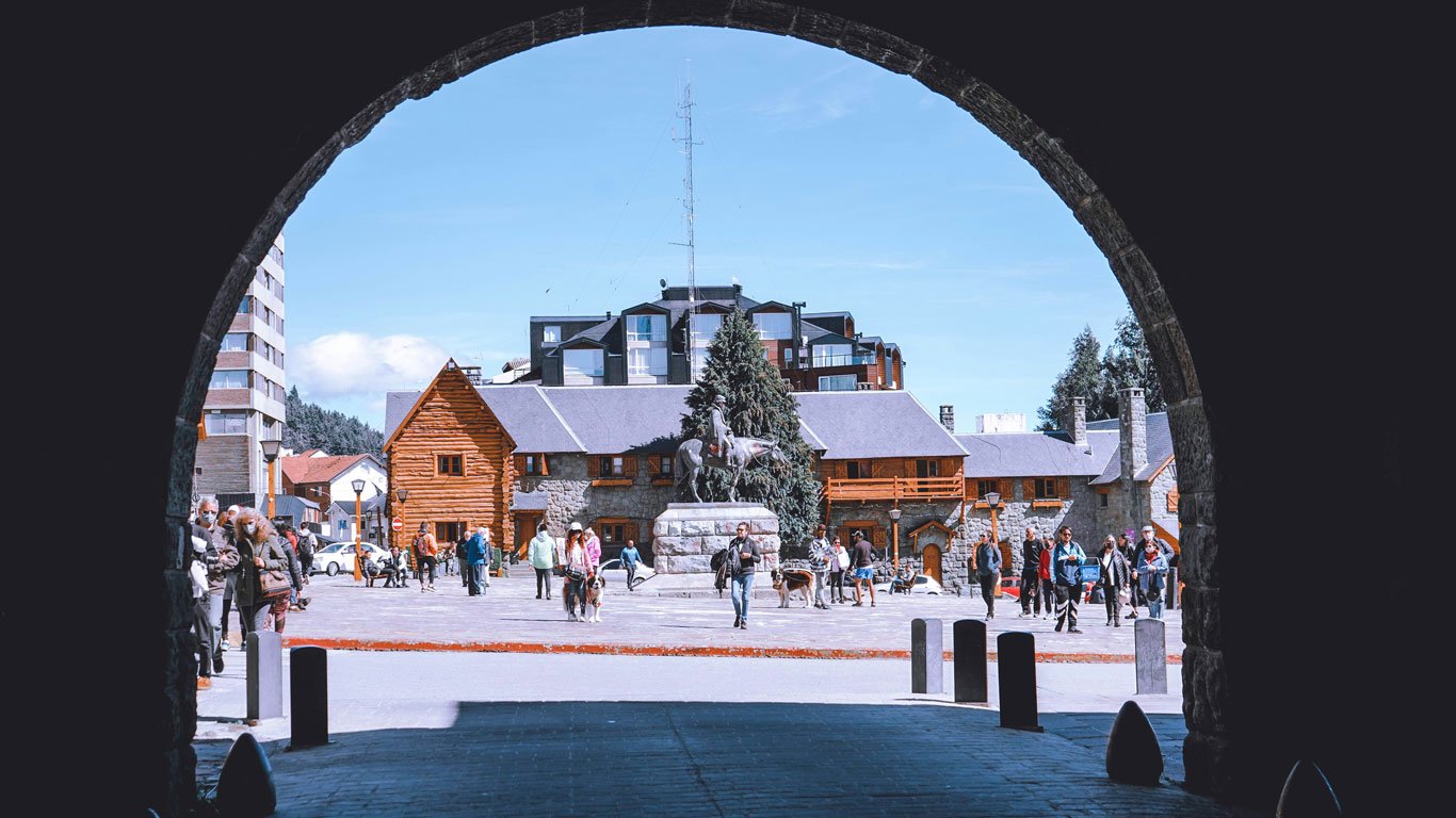 Praça central de Bariloche vista por um arco de pedra, com várias pessoas passeando com roupas de frio, casas de madeira e pedra ao fundo e uma escultura de cavalo no centro. O céu está limpo e azul, revelando um dia frio, porém ensolarado.