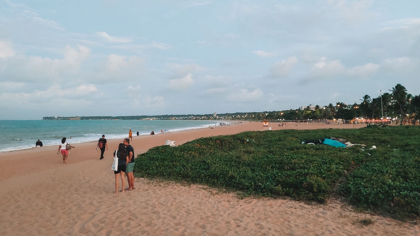 cabo-branco-joao-pessoa Pessoas caminhando e conversando na faixa de areia da Praia de Cabo Branco, com vegetação rasteira à direita e a cidade de João Pessoa ao fundo.