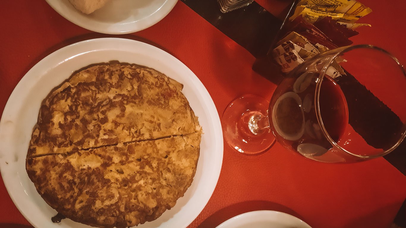 Tortilla de papas, servida em um prato branco sobre uma toalha vermelha, acompanhada de uma taça de vinho.