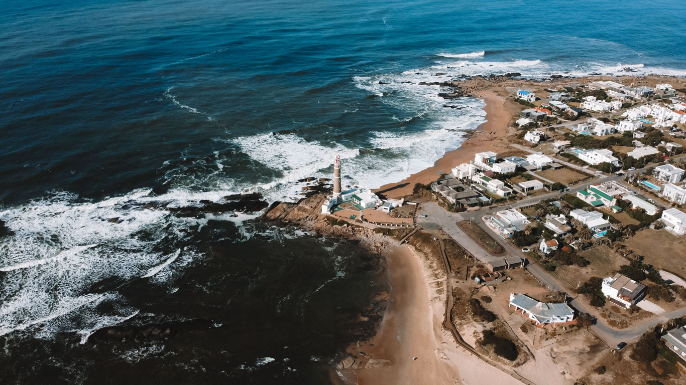 Vista aérea de José Ignacio, Uruguai, mostrando um farol icônico sobre um promontório rochoso cercado por ondas quebrando e praias de areia clara. Ao lado, um pequeno vilarejo costeiro com casas brancas e ruas tranquilas compõem uma paisagem charmosa e sofisticada à beira-mar.