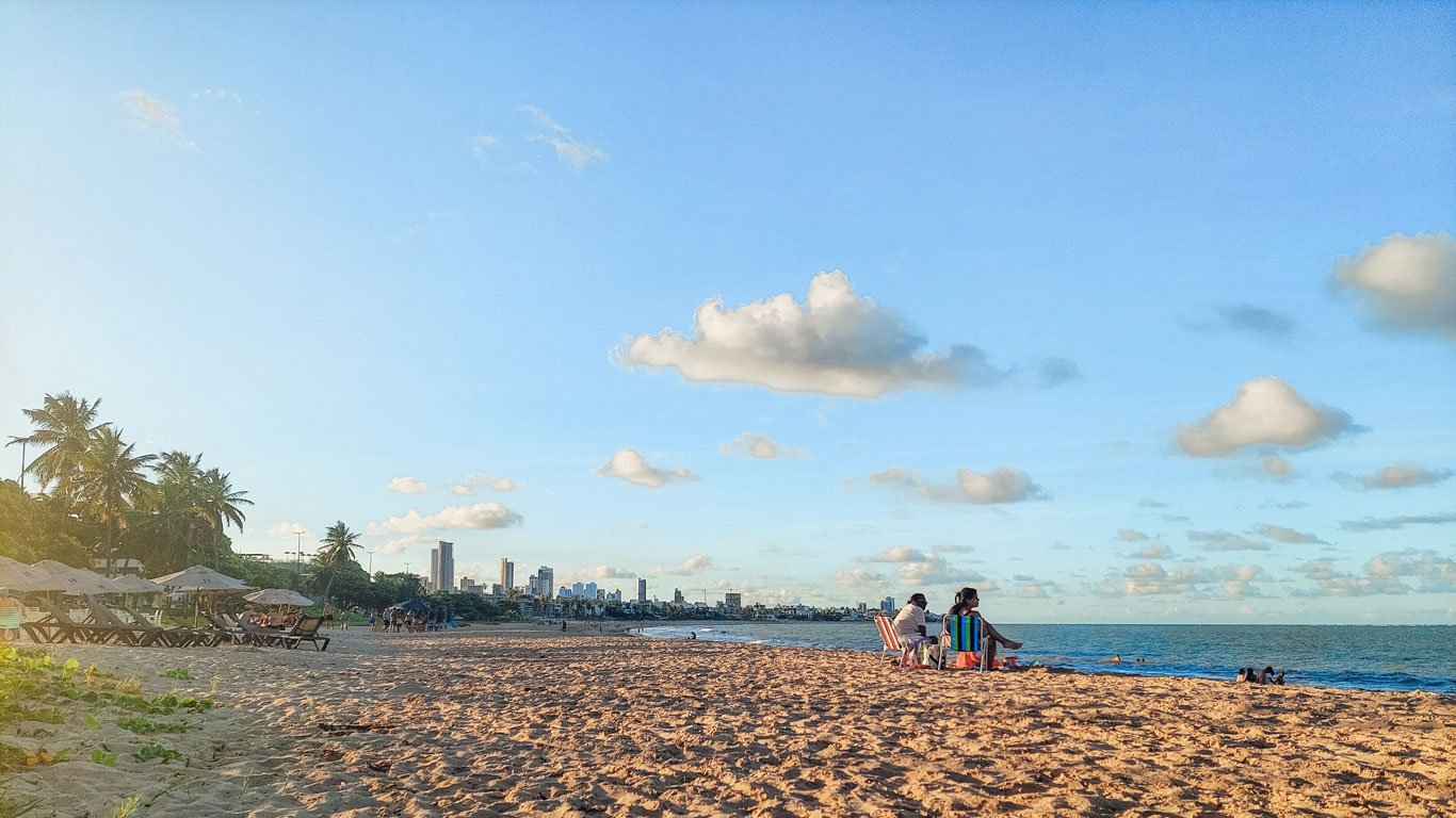 manaira Praia de Manaíra com palmeiras e cadeiras vazias à esquerda, enquanto algumas pessoas relaxam na areia à direita. Ao fundo, o skyline de João Pessoa sob um céu azul dourado do fim da tarde.