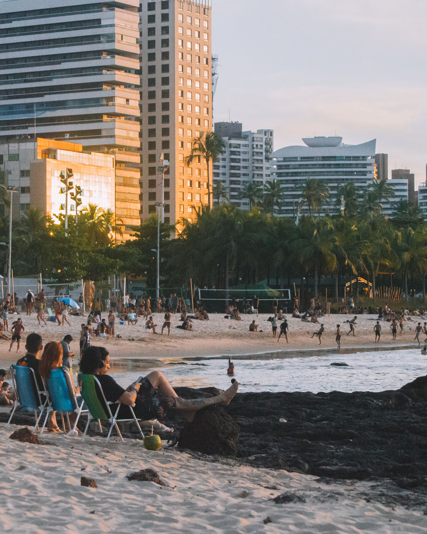 melhor-lugar-para-ficar-em-fortalreza-meireles Multidão aproveitando a Praia de Meireles ao entardecer, o melhor lugar onde ficar em Fortaleza, com palmeiras e edifícios ao fundo, enquanto o sol dourado reflete nos prédios.
