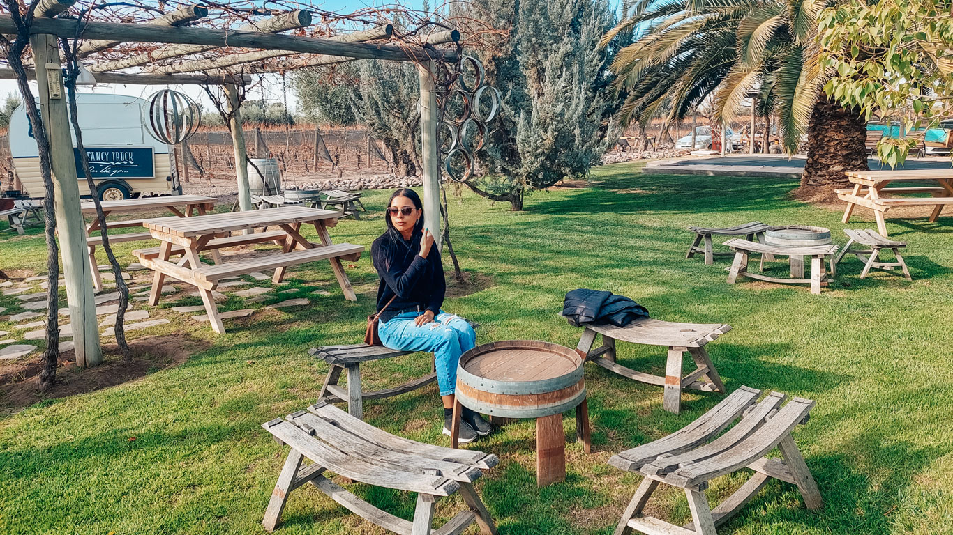 mendoza-em-outubro-1 Mulher sentada em um jardim bem cuidado, na bodega Tempus Alba em Mendoza, rodeada por bancos e mesas de madeira rústica, com um trailer ao fundo.