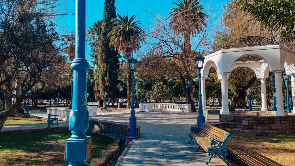 Praça Independência em Mendoza em outubro, arborizada com bancos de madeira, postes azuis e um coreto branco, sob um céu limpo e azul.