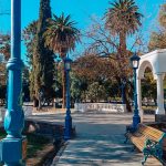 Praça Independência em Mendoza em outubro, arborizada com bancos de madeira, postes azuis e um coreto branco, sob um céu limpo e azul.