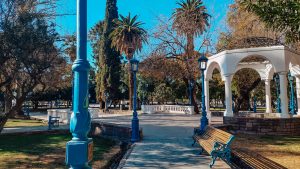 Praça Independência em Mendoza em outubro, arborizada com bancos de madeira, postes azuis e um coreto branco, sob um céu limpo e azul.