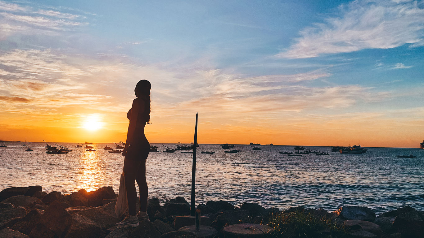 mucuripe-fortaleza Mulher de perfil observando o pôr do sol na orla de Mucuripe, em Fortaleza, com barcos ancorados no mar e o céu pintado de laranja e azul ao fundo.