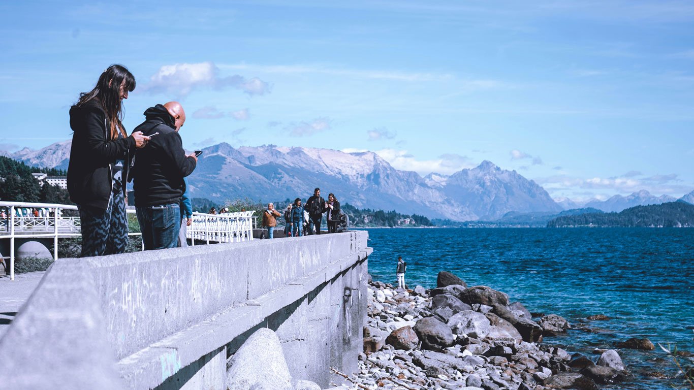 Pessoas caminhando por um píer de concreto à beira do lago Nahuel Huapi em Bariloche, com águas azuis cristalinas, pedras na margem e montanhas ao fundo sob céu claro.