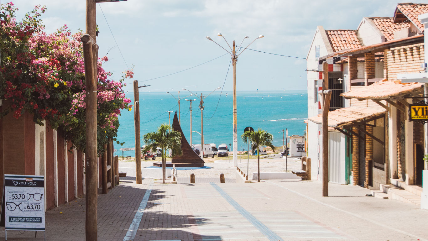 onde-ficar-em-canoa-quebrada Vista da rua Broadway no centro de Canoa Quebrada, que desce em direção ao mar azul, com postes, palmeiras e algumas construções de tijolos e placas comerciais. Uma escultura de vela náutica aparece ao fundo.