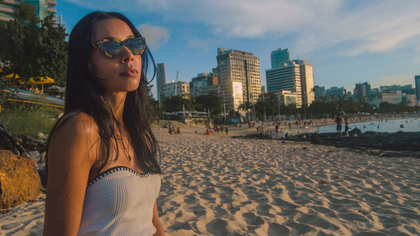 onde-ficar-em-fortaleza-1 Mulher de cabelos longos e óculos de sol posa na areia da Praia de Meireles ao entardecer, com arranha-céus ao fundo refletindo a luz dourada do sol e diversas pessoas aproveitando a praia.