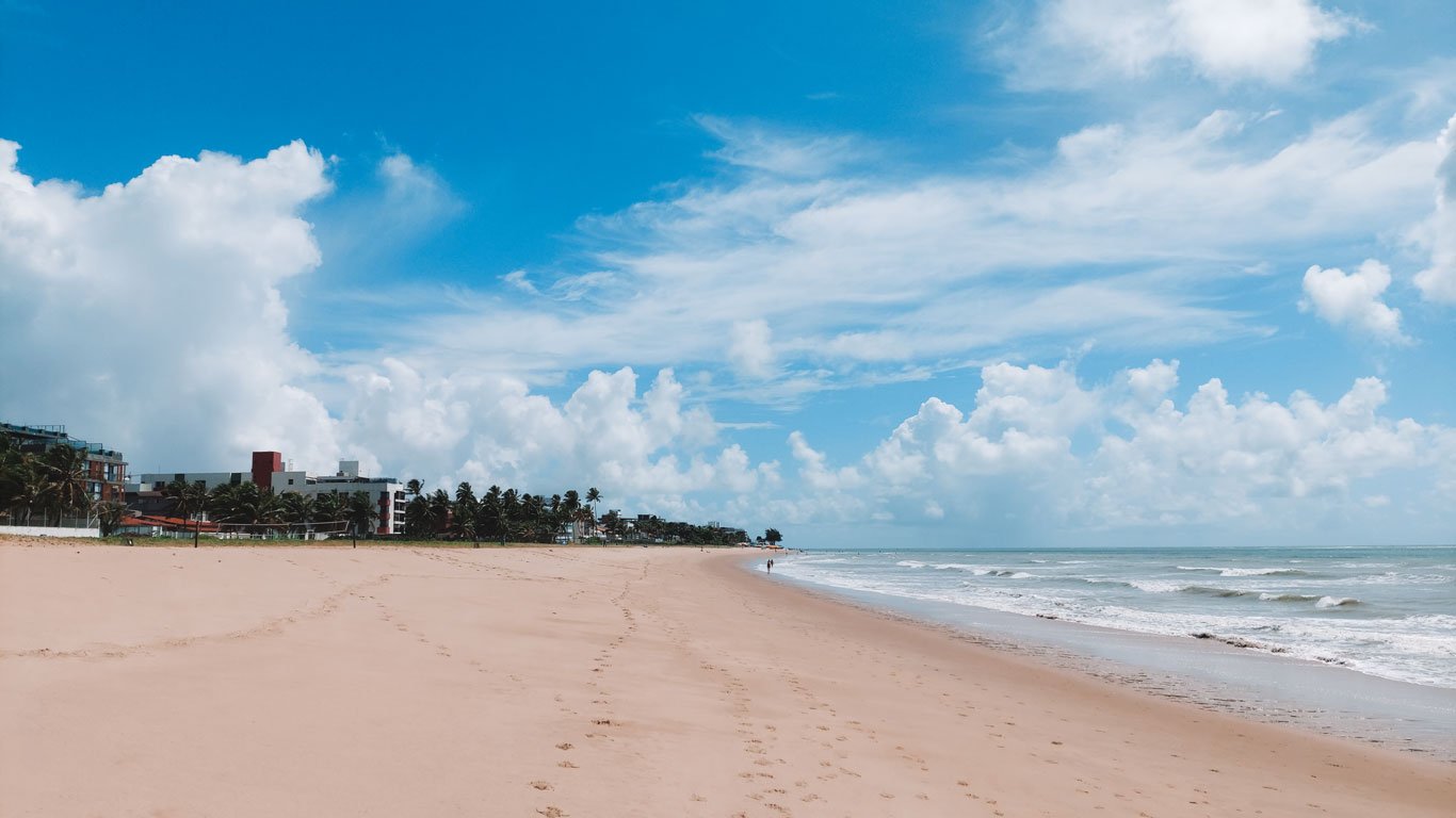 onde-ficar--em-joao-pessoa Praia de Jardim Oceania, sem banhistas, com areia clara e ondas suaves, sob um céu azul com nuvens volumosas. Edifícios baixos e palmeiras à esquerda, em João Pessoa.