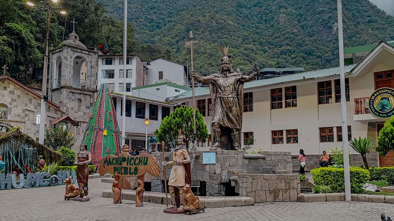 onde-ficar-em-machu-picchu Estátua inca na praça principal de Águas Calientes, rodeada por construções turísticas e vegetação. Esse é o coração de onde ficar em Machu Picchu, com hospedagens, restaurantes e fácil acesso ao ônibus que leva às ruínas.