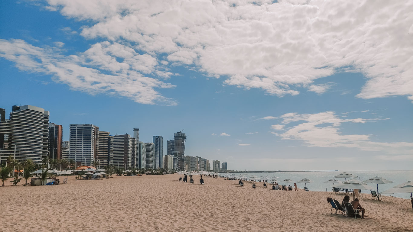 praia-de-iracema-fortaleza Vista ampla da Praia do Iracema em um dia claro, com areia dourada, guarda-sóis brancos e prédios altos à esquerda contrastando com o céu azul pontilhado de nuvens.