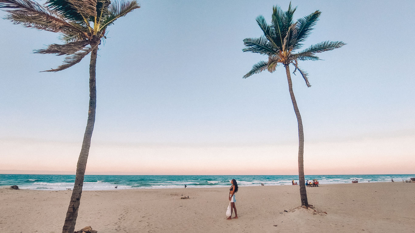 praia-do-futuro Mulher em pé na areia entre duas palmeiras altas na Praia do Futuro, com o mar ao fundo e céu aberto ao entardecer, transmitindo tranquilidade e amplitude.