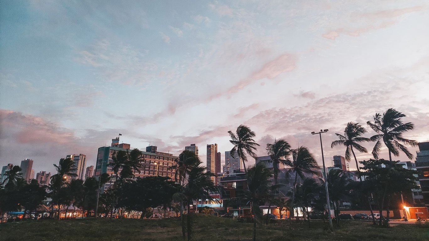 tambau-joao-pessoa Vista urbana de Tambaú em João Pessoa ao pôr do sol, com prédios altos ao fundo e coqueiros balançando à frente. O céu exibe tons rosados e azulados ao entardecer.