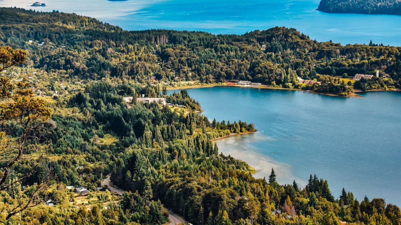 Vista panorâmica do Lago Nahuel Huapi em Bariloche,cercados por florestas densas e pequenas construções espalhadas.