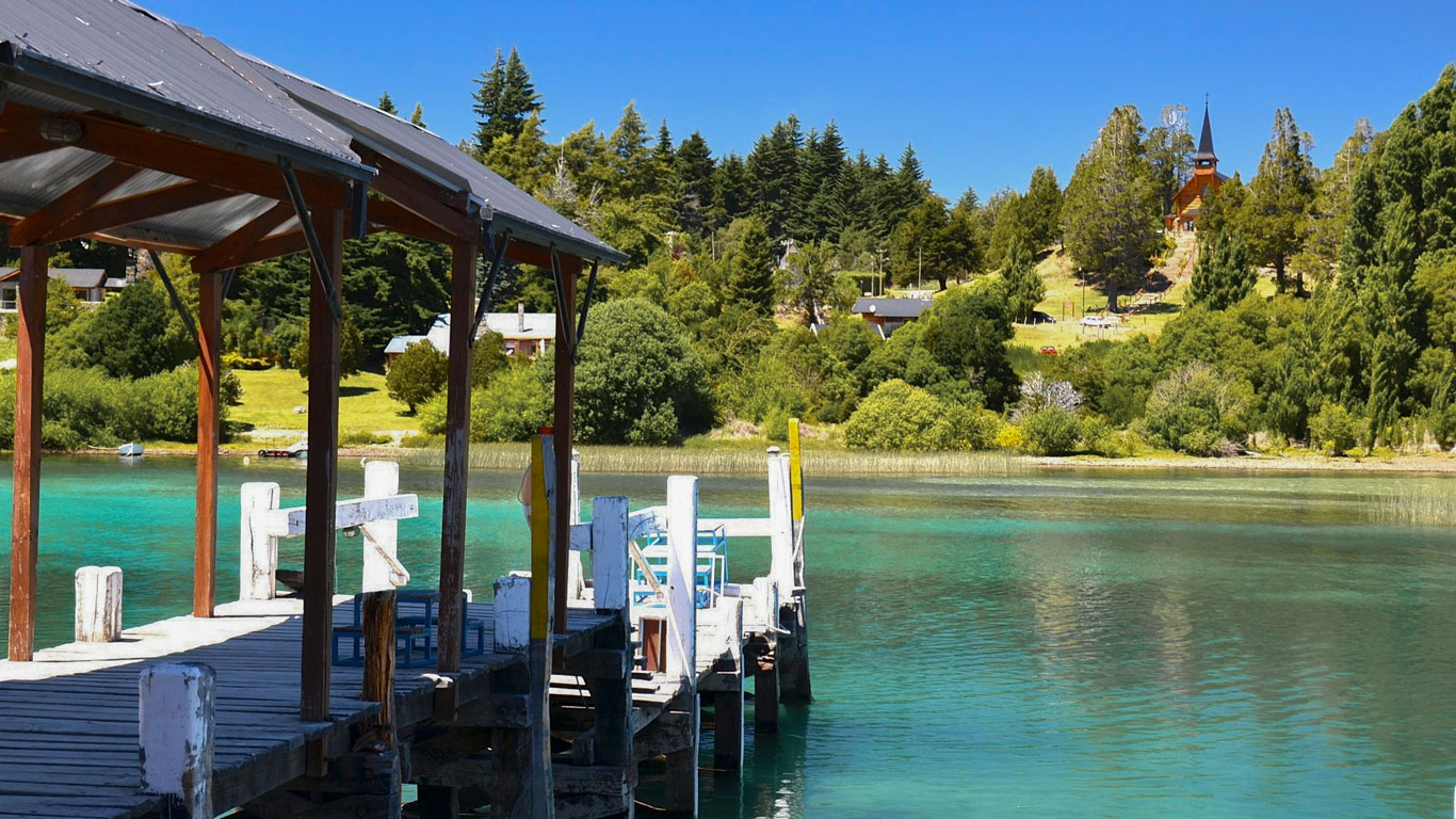 bariloche-em-novembro-1 Cais de madeira antigo à beira de um lago cristalino em Bariloche, com águas verde-esmeralda, rodeado por vegetação exuberante. No alto da colina, uma pequena igreja com torre pontiaguda se destaca entre as árvores.
