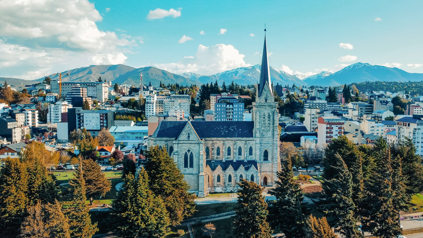 bariloche-em-novembro A Catedral de Bariloche, em estilo gótico, em destaque com seu alto campanário, cercada por árvores e com a cidade e montanhas ao fundo. Céu azul com nuvens e vegetação outonal completam a paisagem urbana.