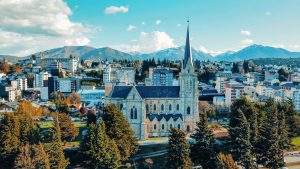 Imagem aérea da cidade de San Carlos de Bariloche, na Argentina, com destaque para a imponente Catedral de Nossa Senhora do Nahuel Huapi em estilo neogótico, rodeada por árvores e edifícios modernos ao fundo. Ao longe, as montanhas cobertas de neve completam a paisagem com um céu azul pontuado por nuvens.