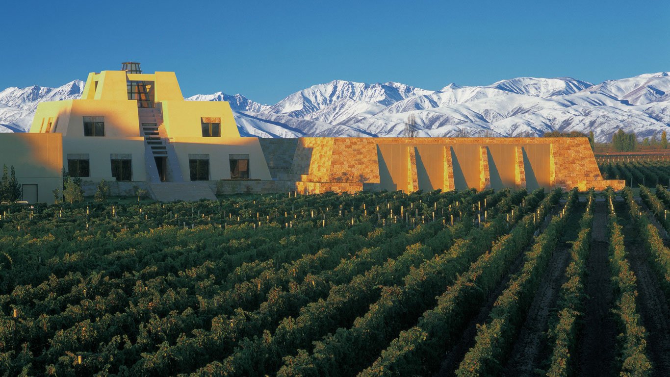 catena-zapata-restaurante Construção moderna da Catena Zapata, com arquitetura piramidal em meio a vinhedos verdes em Mendoza, Argentina, ao entardecer. Ao fundo, a Cordilheira dos Andes aparece coberta de neve sob um céu azul claro, criando um contraste impressionante com a paisagem.