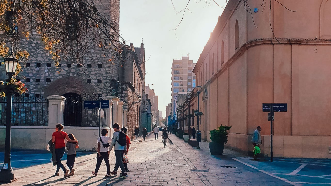 centro-de-cordoba-argentina Pessoas caminhando em uma rua de paralelepípedos no centro histórico de Córdoba, Argentina, ao entardecer. À esquerda, um prédio de pedra antiga e à direita, construções coloniais iluminadas pela luz dourada do sol.