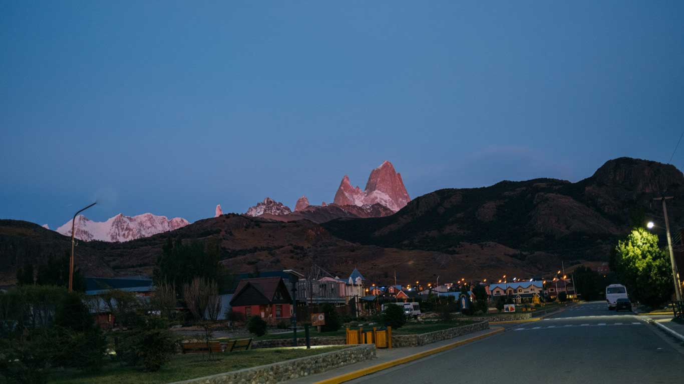 el-chalten Vista ao entardecer da cidade de El Chaltén, com o céu azul escurecendo e as montanhas cobertas de neve ao fundo, incluindo o icônico Monte Fitz Roy iluminado pela luz rosada do pôr do sol.