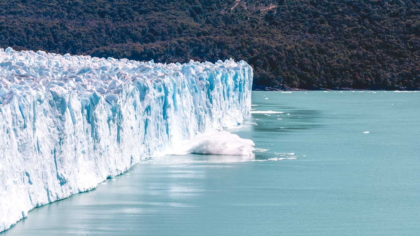 excursao-glaciar-perito-moreno-el-calafate Bloco de gelo gigante se desprendendo do Glaciar Perito Moreno, um dos melhores passeios em El Calafate, e caindo no Lago Argentino, com água salpicando e floresta ao fundo.