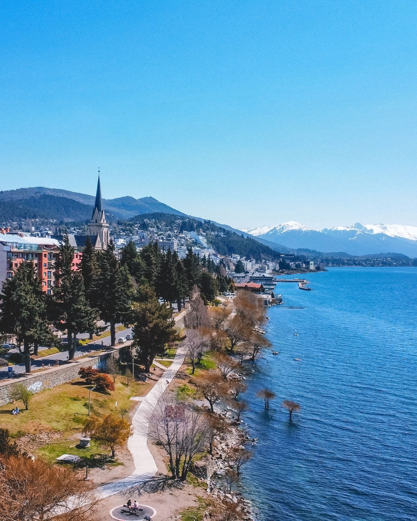 Vista do centro de Bariloche, mostrando edifícios coloridos, uma igreja com torre alta e um calçadão à beira do Lago Nahuel Huapi com montanhas nevadas ao fundo.